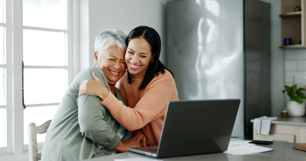 A senior woman with short gray hair and a younger woman with long dark hair share a warm embrace while smiling. They are sitting in a bright kitchen with a laptop open on the counter to personal care communities, surrounded by papers. Natural light streams in through large windows, creating a cozy and happy atmosphere.